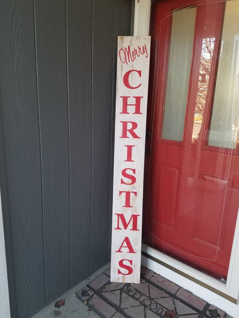 Puede incluir: Un letrero de madera blanco con las palabras "Merry Christmas" en letras rojas. El letrero est&aacute; apoyado contra una puerta roja.