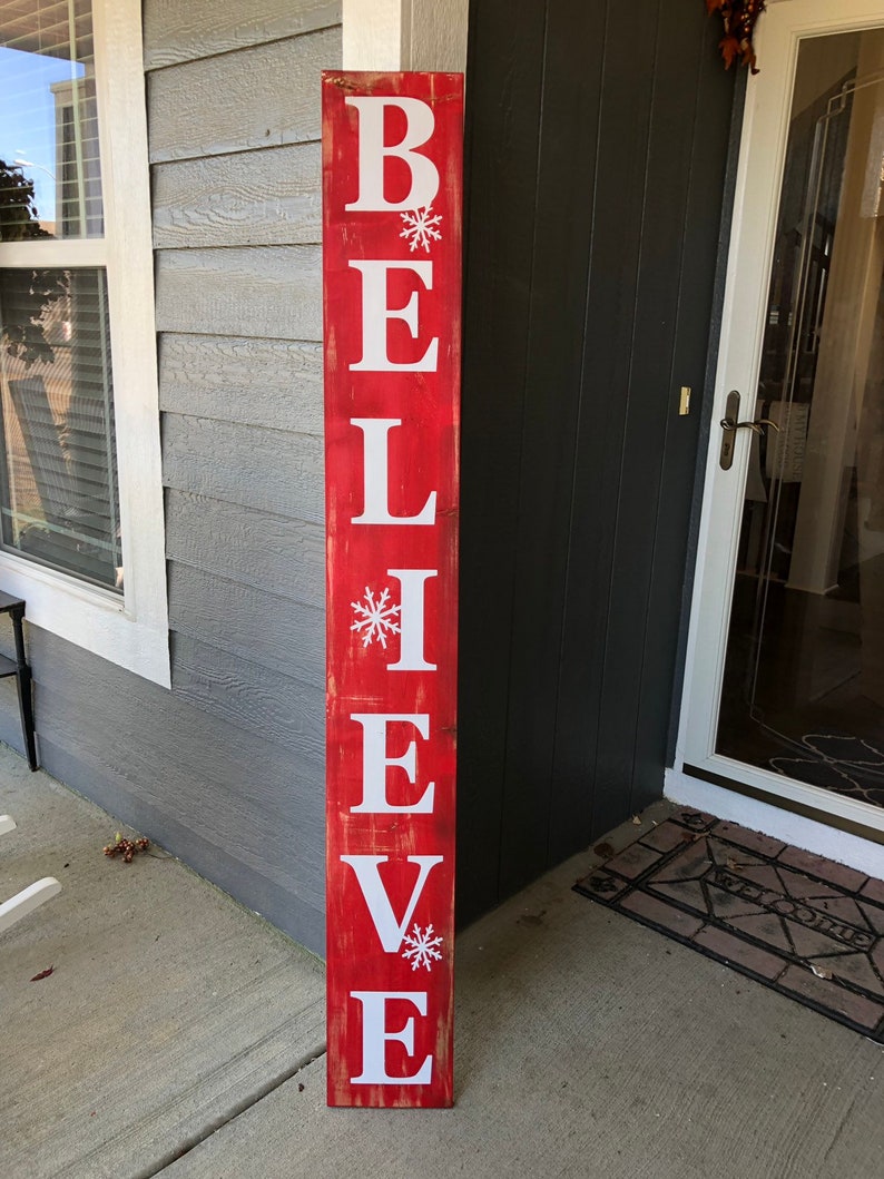 Puede incluir: Un letrero de madera rojo con la palabra "BELIEVE" en letras blancas con un copo de nieve entre cada letra.