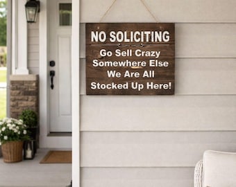 May include: A rectangular wooden sign with the words "NO SOLICITING" in white lettering. Below, the sign humorously states, "Go Sell Crazy Somewhere Else We Are All Stocked Up Here!" The sign hangs on a light gray wall.