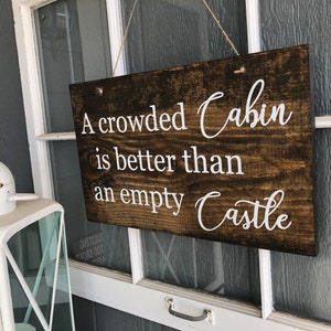 May include: A wooden sign with white text that reads "A crowded Cabin is better than an empty Castle".