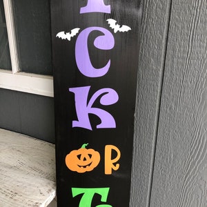 May include: A black wooden sign with the words "Trick or Treat" in purple, orange, and green letters. There are white bats flying above the word "Trick".