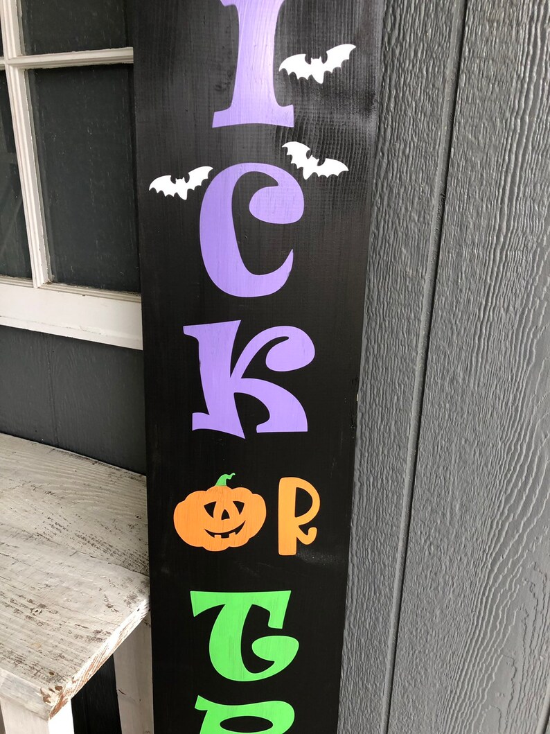 May include: A black wooden sign with the words "Trick or Treat" in purple, orange, and green letters. There are white bats flying above the word "Trick".