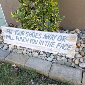 Pu&ograve; includere: Un cartello in legno rustico con il testo "PUT YOUR SHOES AWAY OR I WILL PUNCH YOU IN THE FACE Love, MOM" in vernice blu.