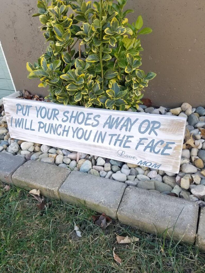 Pu&ograve; includere: Un cartello in legno rustico con il testo "PUT YOUR SHOES AWAY OR I WILL PUNCH YOU IN THE FACE Love, MOM" in vernice blu.