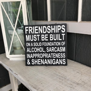 May include: Black wooden sign with white text that reads "Friendships must be built on a solid foundation of alcohol, sarcasm, inappropriateness & shenanigans."