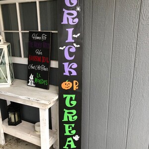 May include: Black wooden sign with the words "Trick or Treat" in green and purple letters. The sign has a spider web, spider, bats, a pumpkin, and a ghost.