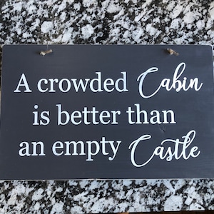 May include: A dark gray wooden sign with white text that reads "A crowded Cabin is better than an empty Castle".