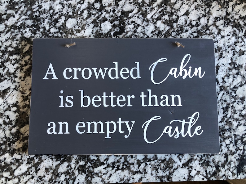 May include: A dark gray wooden sign with white text that reads "A crowded Cabin is better than an empty Castle".