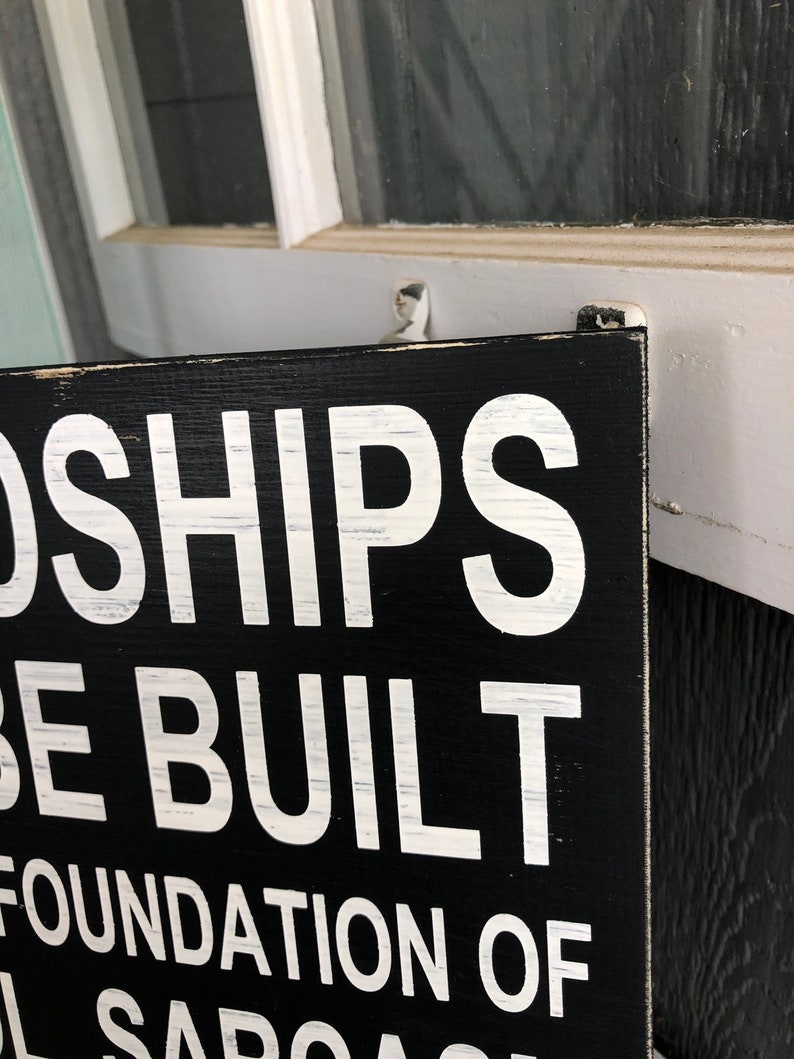 May include: Black wooden sign with white text that reads "Friendships are built on a foundation of mutual sarcasm".