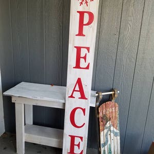 Puede incluir: Un letrero de madera blanco con la palabra "PAZ" en letras rojas. El letrero est&aacute; decorado con copos de nieve rojos en la parte superior e inferior. El letrero est&aacute; apoyado en una mesa de madera blanca. Un trineo rojo y verde con la palabra "Bienvenido" pintada en &eacute;l est&aacute; en el fondo.
