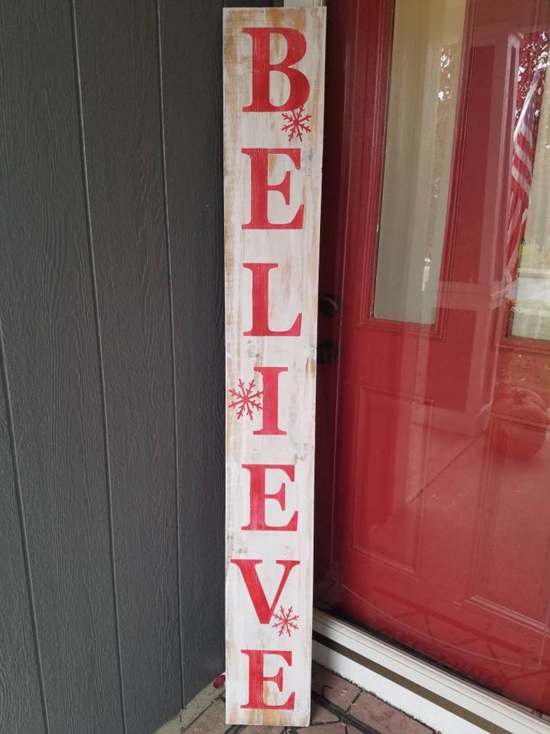 Puede incluir: Un letrero de madera blanco con la palabra "BELIEVE" pintada en letras rojas. Hay peque&ntilde;as estrellas de nieve rojas pintadas entre las letras.
