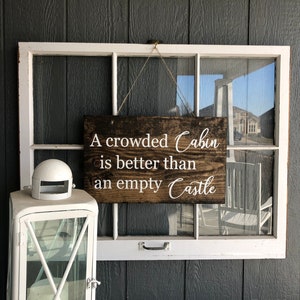 May include: A wooden sign with white text that reads "A crowded Cabin is better than an empty Castle". The sign is hanging from a window frame.