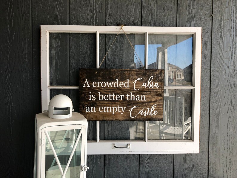 May include: A wooden sign with white text that reads "A crowded Cabin is better than an empty Castle". The sign is hanging from a window frame.