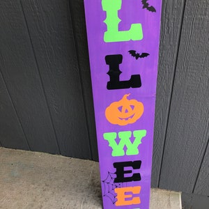 May include: Purple wooden sign with the word "HALLOWEEN" spelled out in black, green, and orange letters. The letters are in a vintage style font. There are black bats and a spider web on the sign.