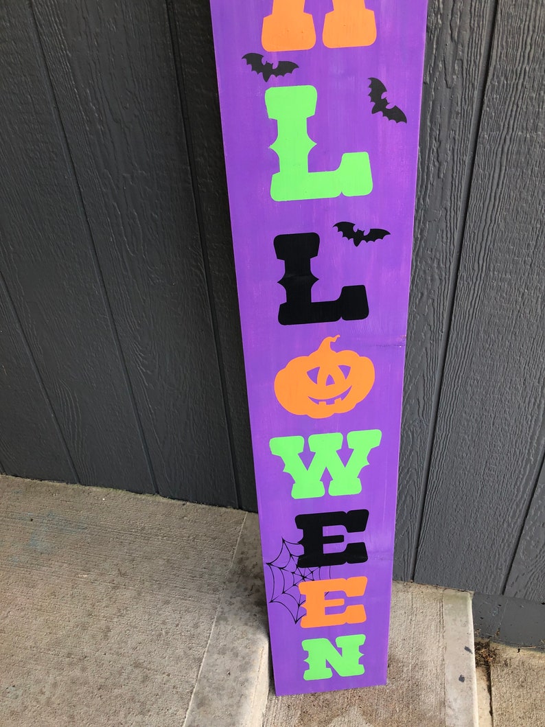 May include: Purple wooden sign with the word "HALLOWEEN" spelled out in black, green, and orange letters. The letters are in a vintage style font. There are black bats and a spider web on the sign.