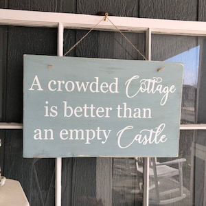 May include: A blue wooden sign with white lettering that reads "A crowded Cottage is better than an empty Castle".