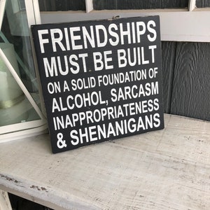 May include: Black wooden sign with white text that reads "Friendships must be built on a solid foundation of alcohol, sarcasm, inappropriateness & shenanigans."