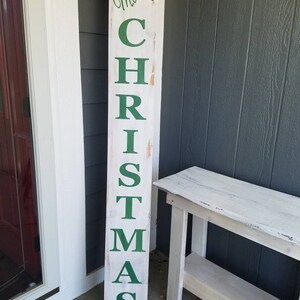 Puede incluir: Un letrero de madera blanco con las palabras "Merry Christmas" en letras verdes. El letrero est&aacute; apoyado en una mesa de madera blanca.