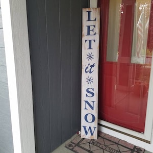 Puede incluir: Un letrero de madera blanco con letras azules que dice "LET it SNOW" con un dise&ntilde;o de copo de nieve arriba y debajo de la palabra "it". El letrero est&aacute; apoyado contra una casa gris con una puerta roja.
