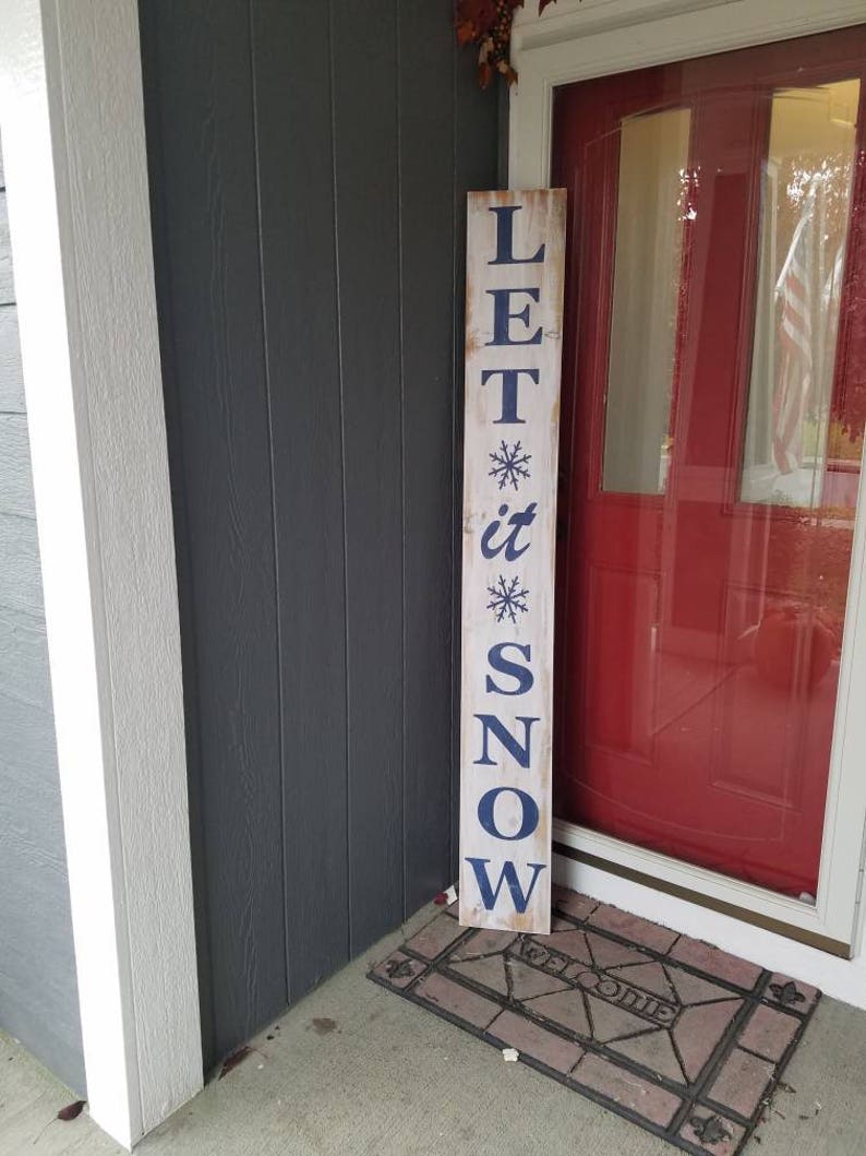 Puede incluir: Un letrero de madera blanco con letras azules que dice "LET it SNOW" con un dise&ntilde;o de copo de nieve arriba y debajo de la palabra "it". El letrero est&aacute; apoyado contra una casa gris con una puerta roja.