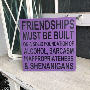 May include: Purple wooden sign with white lettering that reads "Friendships must be built on a solid foundation of alcohol, sarcasm, inappropriateness & shenanigans."