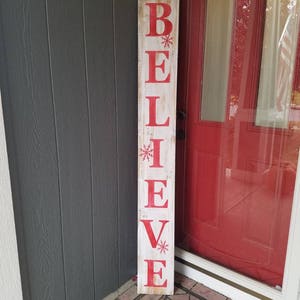 Puede incluir: Un letrero de madera blanco con la palabra "BELIEVE" pintada en letras rojas. El letrero tiene un aspecto desgastado y est&aacute; decorado con peque&ntilde;as estrellas de nieve rojas.