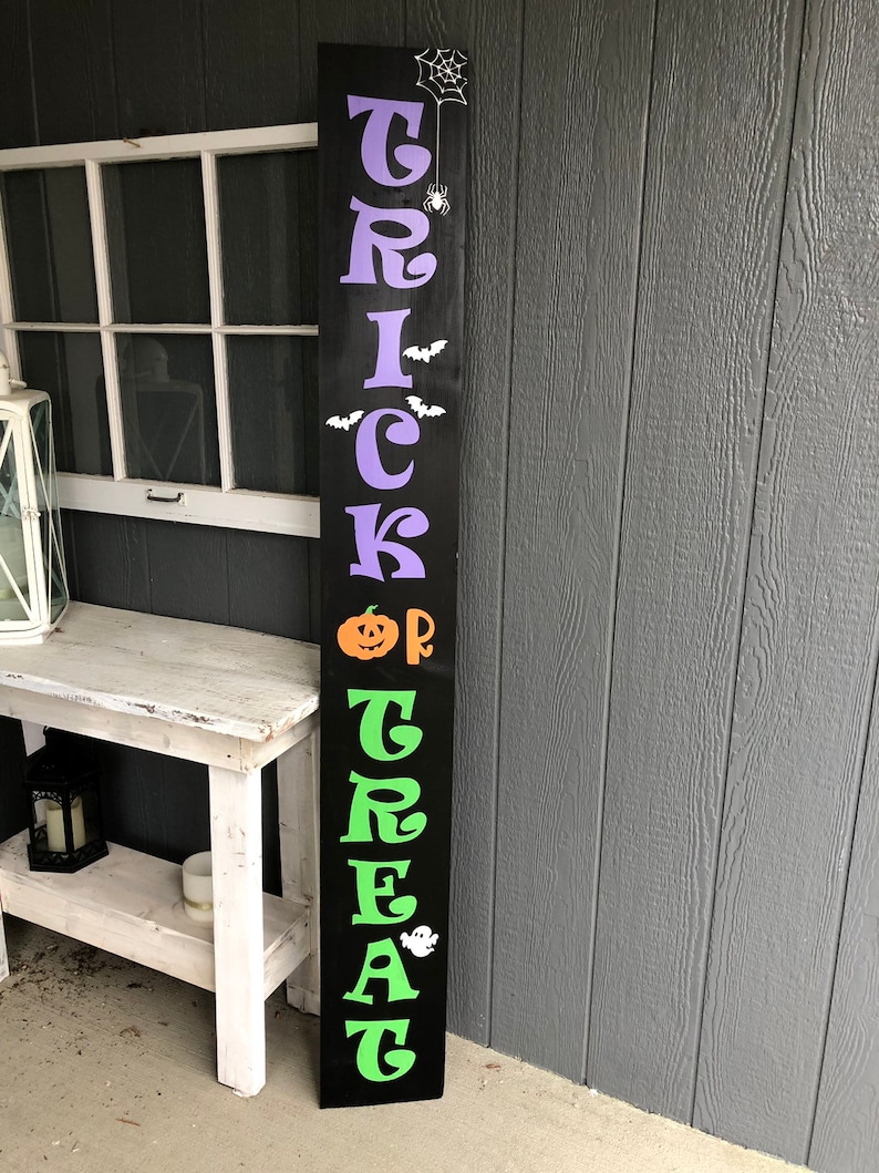 May include: A black wooden sign with the words "Trick or Treat" in green and purple letters. The sign also features a pumpkin, a spider, bats, and a ghost.