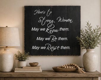 Puede incluir: Un letrero cuadrado de madera negra con un acabado desgastado que presenta texto blanco en cursiva que dice: Here's to Strong Women, May we know them. May we be them. May we raise them. El letrero está hecho de tablones verticales y tiene una textura mate.