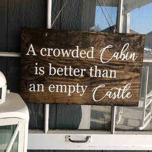 May include: A wooden sign with a dark brown stain hangs on a white door. The sign reads "A crowded Cabin is better than an empty Castle" in white lettering.