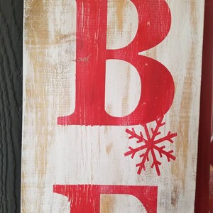 Puede incluir: Un letrero de madera con las letras "BE" pintadas de rojo. Un copo de nieve rojo est&aacute; pintado entre las letras.
