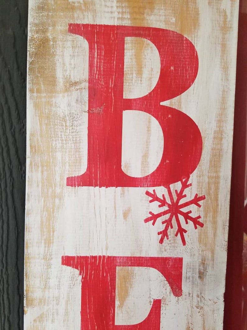 Puede incluir: Un letrero de madera con las letras "BE" pintadas de rojo. Un copo de nieve rojo est&aacute; pintado entre las letras.