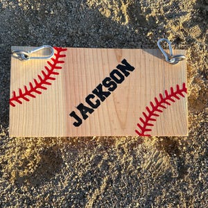 May include: A wooden sign with a baseball stitch design and the name "JACKSON" in black lettering. The sign has two metal hooks on the top.