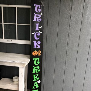 May include: Black wooden sign with the words "Trick or Treat" in purple, green, and orange letters. The sign has a spider web and spider, bats, a pumpkin, and a ghost.