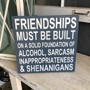 May include: A dark grey wooden sign with white text that reads "Friendships must be built on a solid foundation of alcohol, sarcasm, inappropriateness & shenanigans."