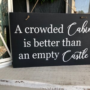 May include: A black wooden sign with white text that reads "A crowded Cabin is better than an empty Castle".