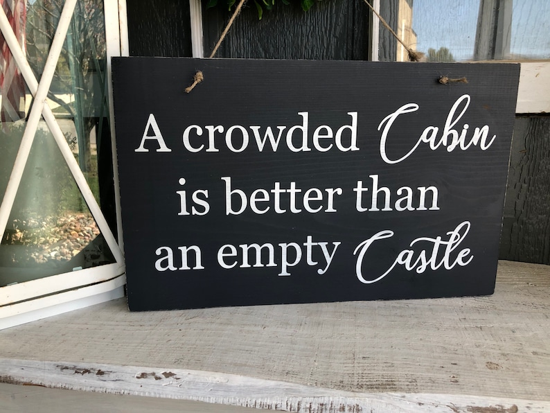 May include: A black wooden sign with white text that reads "A crowded Cabin is better than an empty Castle".