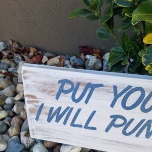 Pu&ograve; includere: Un cartello di legno bianco con testo dipinto in blu che dice "Put your  I will pun". Il cartello &egrave; appoggiato su un letto di piccole pietre colorate.