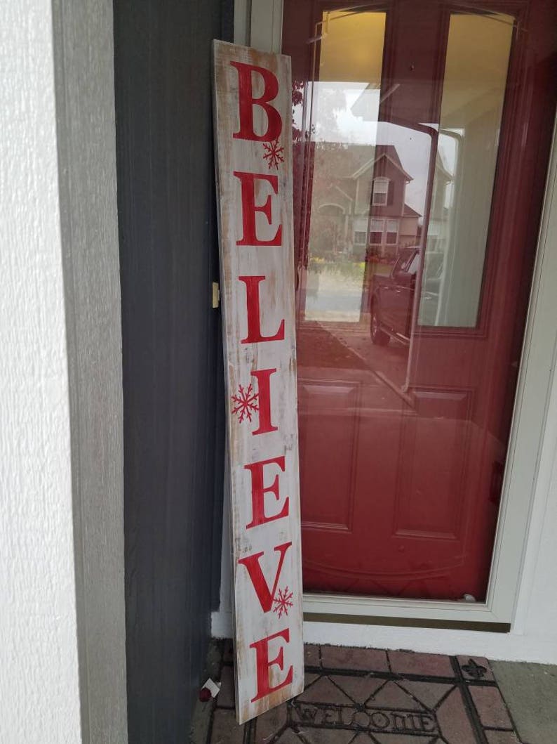 Puede incluir: Un letrero de madera con la palabra "BELIEVE" pintada en letras rojas con copos de nieve. El letrero est&aacute; desgastado y tiene un aspecto r&uacute;stico.