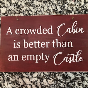 May include: A red wooden sign with white text that reads "A crowded Cabin is better than an empty Castle".