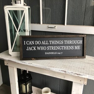 Op de afbeelding: Een zwart ingelijst bord met witte tekst die luidt: "Ik kan alles doen door Jack die mij sterkt. Dani&euml;l 24:7". Het bord staat op een witte houten tafel.