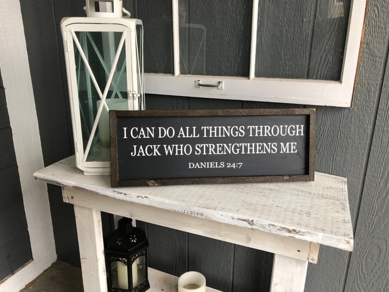 Op de afbeelding: Een zwart ingelijst bord met witte tekst die luidt: "Ik kan alles doen door Jack die mij sterkt. Dani&euml;l 24:7". Het bord staat op een witte houten tafel.