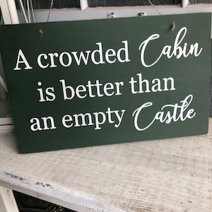 May include: A green wooden sign with white text that reads "A crowded Cabin is better than an empty Castle".