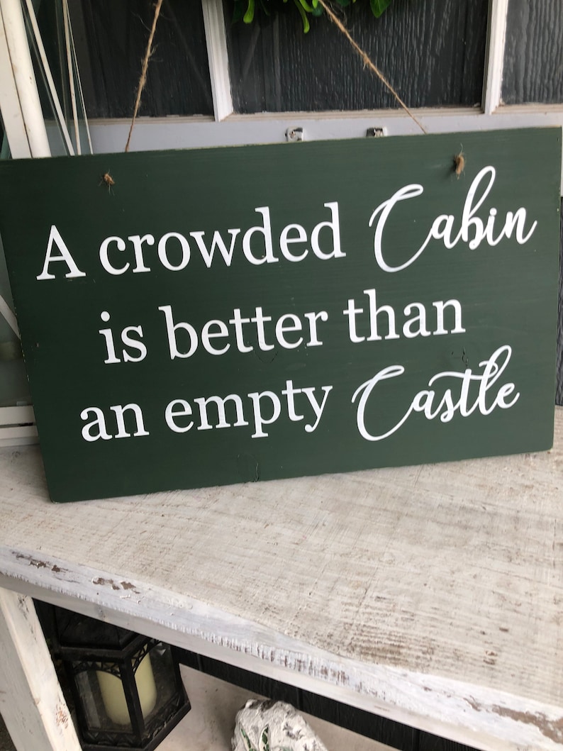 May include: A green wooden sign with white text that reads "A crowded Cabin is better than an empty Castle".
