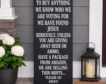 May include: A black rectangular sign with white text that reads "NO SOLICITING...PLEASE GO AWAY!" The sign is displayed on a porch, next to a potted plant with white flowers and a black lantern with a lit candle.