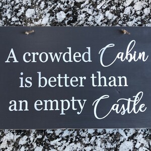 May include: A dark gray wooden sign with white text that reads "A crowded Cabin is better than an empty Castle".