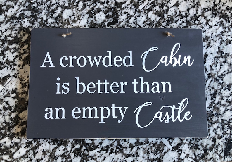 May include: A dark gray wooden sign with white text that reads "A crowded Cabin is better than an empty Castle".