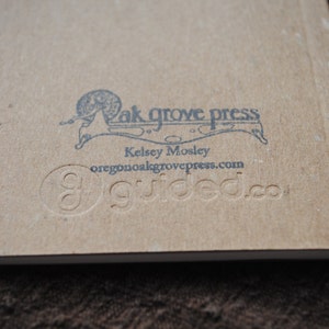 May include: A brown paper book with a black and white illustration of a snail on the back cover. The text "Oak Grove Press" is printed above the snail illustration. The text "Kelsey Mosley" is printed below the snail illustration. The text "oregon@oakgrovepress.com" is printed below the name. The text "guided.co" is printed below the email address.
