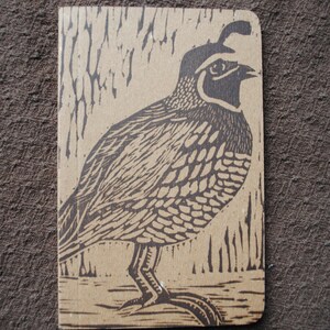 May include: A brown and black linocut print of a quail on a brown background. The quail is facing right and has a detailed pattern of feathers.