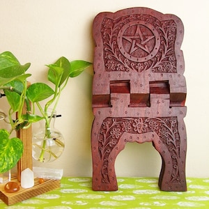 Puede incluir: Un soporte de libro de madera de color rojo oscuro con intrincados tallados de flores y una estrella de cinco puntas. El soporte está diseñado para sostener un libro abierto en ángulo.