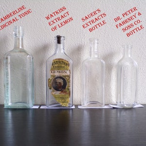 May include: Four clear glass bottles with labels. The bottles are lined up on a dark wooden surface. The labels read "Chamberline Medicinal Tonic", "Watkins Extract of Lemon", "Sauer's Extracts Bottle", and "Dr. Peter Fahrney & Sons Co. Bottle".